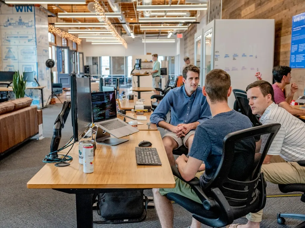 Three colleagues collaborating at a desk with a laptop showing code in a modern open-plan office